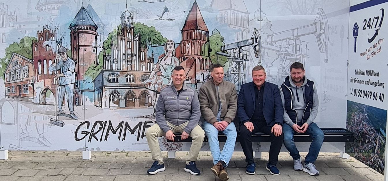 Bürgermeister Marco Jahns, Künstler Christoph Kadur, GWG Technikleiter Björn Wodzich und Werbefachmann Reno Zülsdorf sitzen in der neu gestalteten Busbude am Bahnhof. Im Hintergrund, ist die neue Graftik der Stadt Grimmen zu sehen.