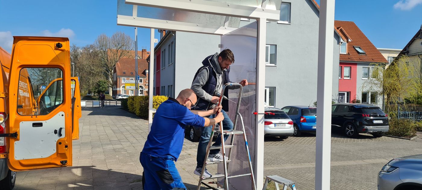 Zwei Arbeiter montieren die neuen, mit der Grafik bedruckten Rückwände an der Bushaltestelle am Bahnhof.