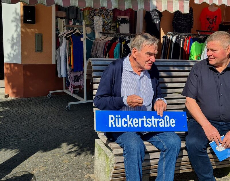 Axel Rückert sitzt mit Bürgermeister Marco Jahns auf einer Bank auf dem Marktplatz. Sie unterhalten sich. Axel Rückert hält ein Straßenschild in der Hand. Es trägt den Namenszug seiner Familie.