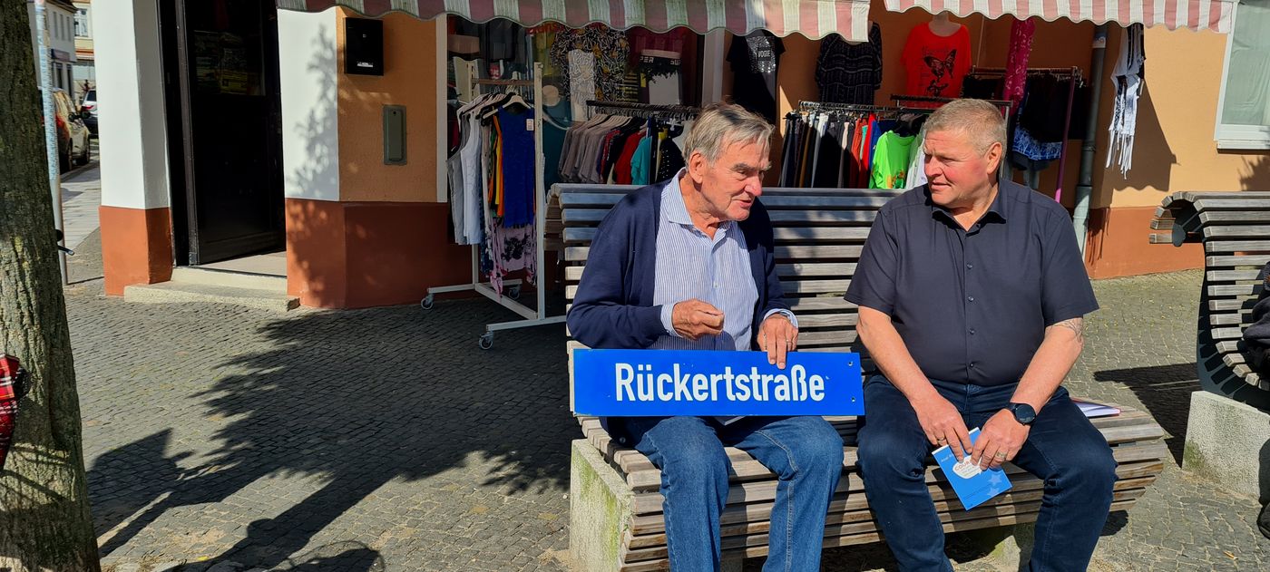 Axel Rückert sitzt mit Bürgermeister Marco Jahns auf einer Bank auf dem Marktplatz. Sie unterhalten sich. Axel Rückert hält ein Straßenschild in der Hand. Es trägt den Namenszug seiner Familie.