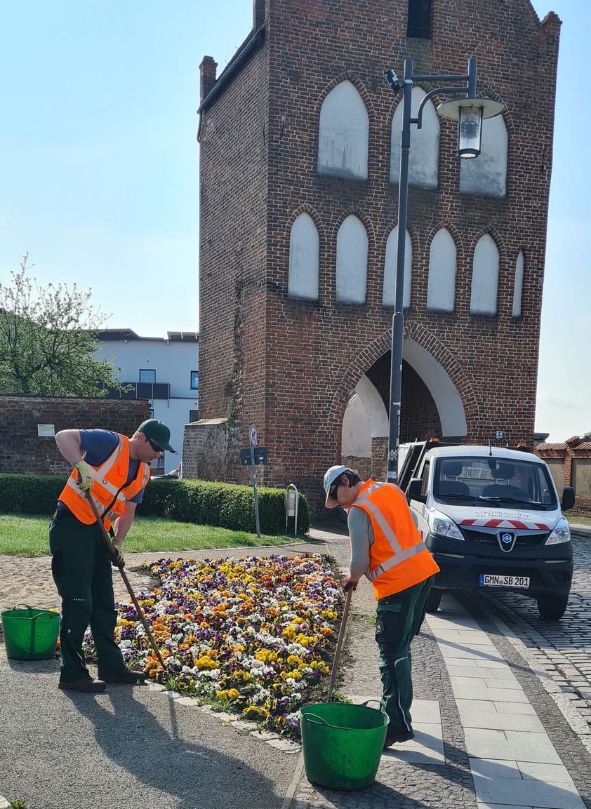 Das Foto zeigt zwei Mitarbeiter des Bauhofes bei der Pflege eines Blumenbeetes vor dem Stadttor.