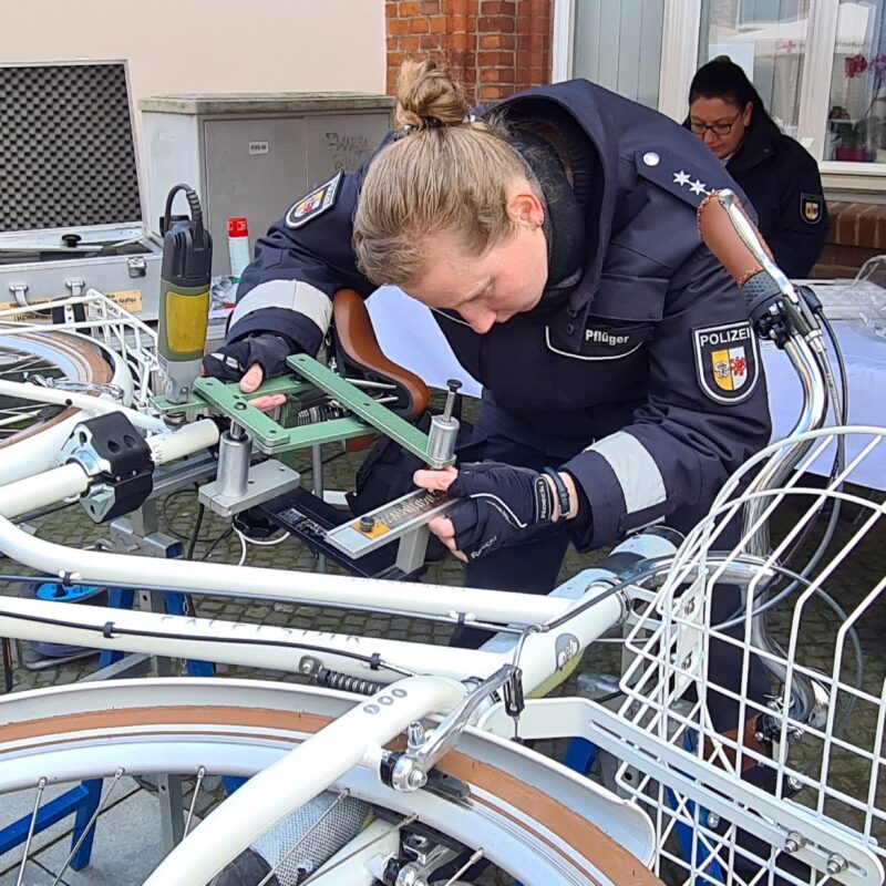 Polizistin beugt sich über ein auf der Seite liegendes Fahrrad. Mit einer Fräse codiert sie den Fahrradrahmen.