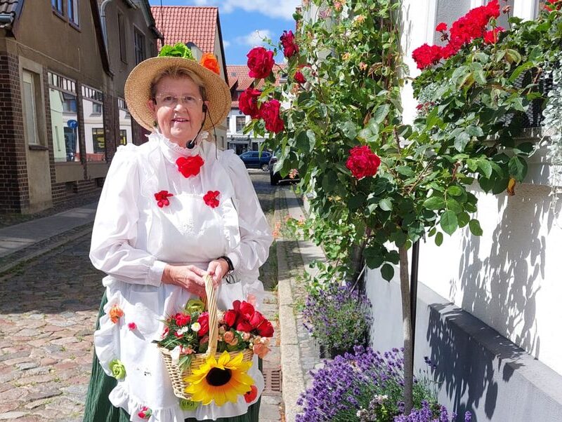 Stadtführerin Gudrun Rech posiert, als Blumenfrau verkleidet, vor einem historischen Gebäude der Stadt. In der Hand hält sie einen Korb mit Sonnenblumen.