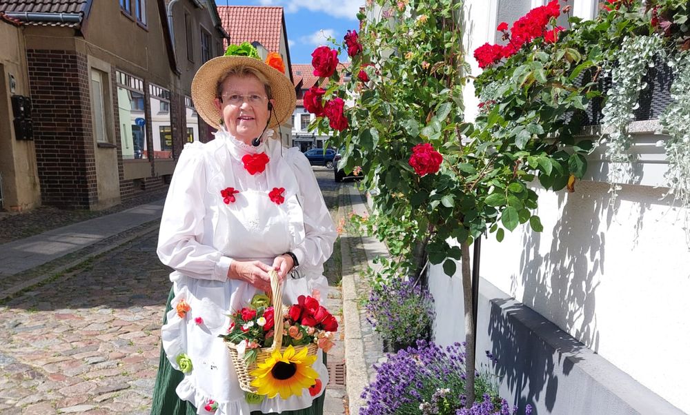 Stadtführerin Gudrun Rech posiert, als Blumenfrau verkleidet, vor einem historischen Gebäude der Stadt. In der Hand hält sie einen Korb mit Sonnenblumen.
