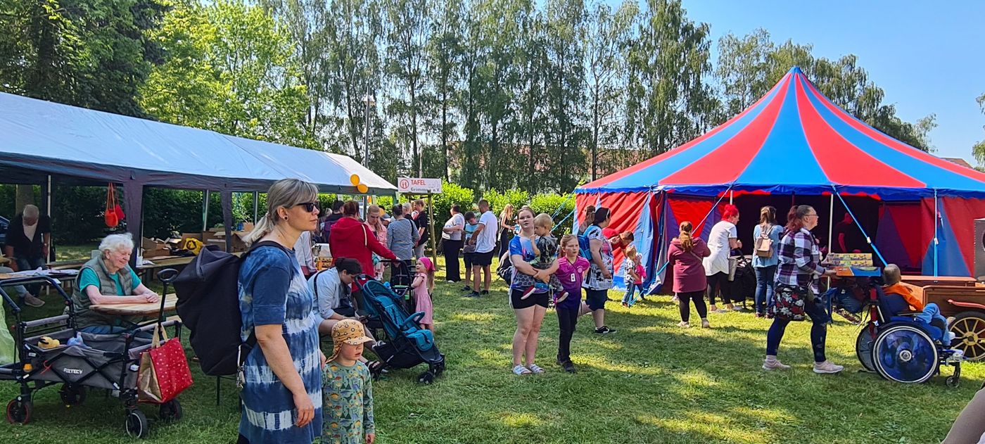 Die Menschen genießen einen Jahrmarkt im Freien mit einem rot-blauen Zirkuszelt, Essensständen und Tischen unter einem Vordach; Familien, Kinder und ältere Menschen mischen sich an einem sonnigen Tag mit Bäumen im Hintergrund.