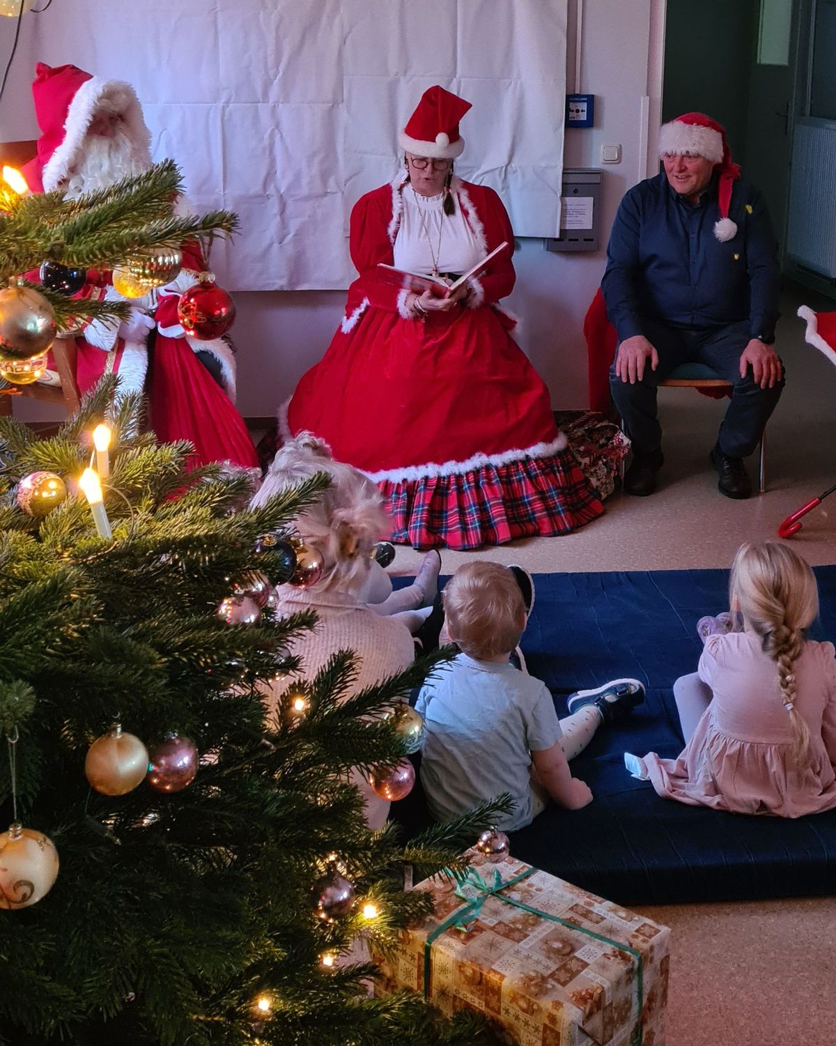 In dieser Kita sitzen die Kinder auf einer Decke vor dem Weihnachtsmann und lauschen seinen Worten. Im Vordergrund, ist ein liebevoll geschmückter Weihnachtsbaum zu sehen.