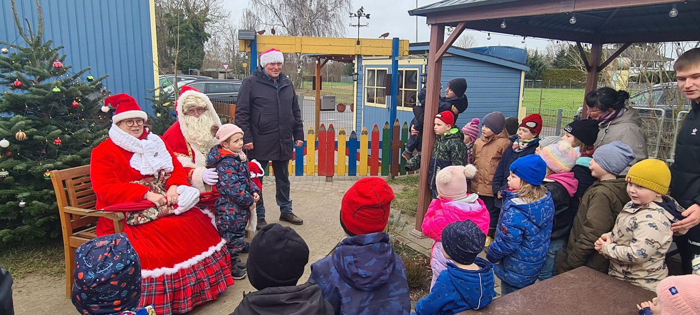 Der Weihnachtsmann und die Märchentante sitzen auf einer bank. Der Bürgermeister steht daneben. Die Kinder blicken gespannt zu den dreien und lauschen einer Geschichte.