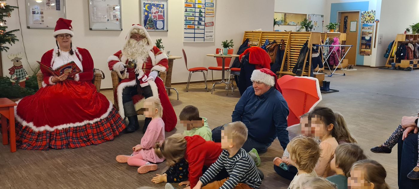 Die Kinder sitzen in der Kita auf dem Fußboden. Auch der Bürgermeister. neben dem tannenbaum sitzen Nicolina und der Weihnachtsmann. Märchentante Nicolina liest eine Geschichte vor. Die Kinder hören gespannt zu.