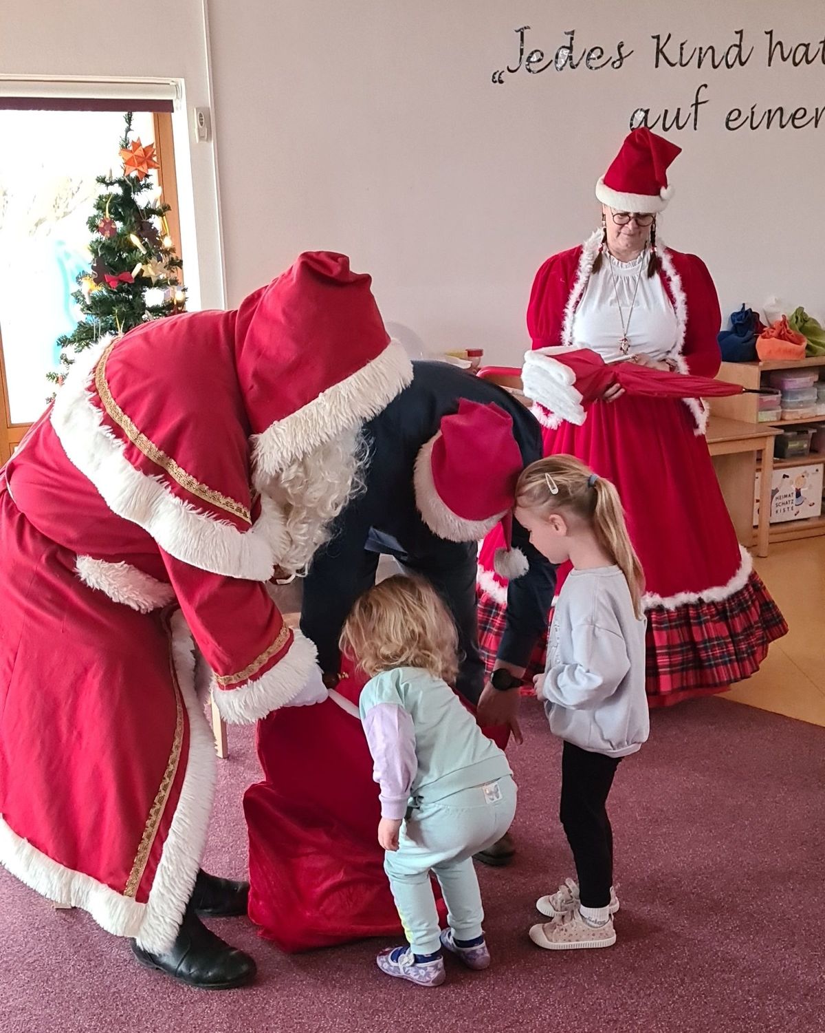 Der Weihnachtsmann und der Bürgermeister beschenken zwei kleine Mädchen. Die schauen gespannt in den Geschenkesack.