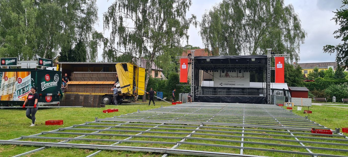 Arbeiter bauen eine Bühne auf und verlegen Metallträger für den Bodenbelag auf einer Wiese. Ein Getränkewagen und ein gelber Lkw mit Ausrüstung sind in der Nähe geparkt. Im Hintergrund sind hohe Bäume und Gebäude zu sehen.