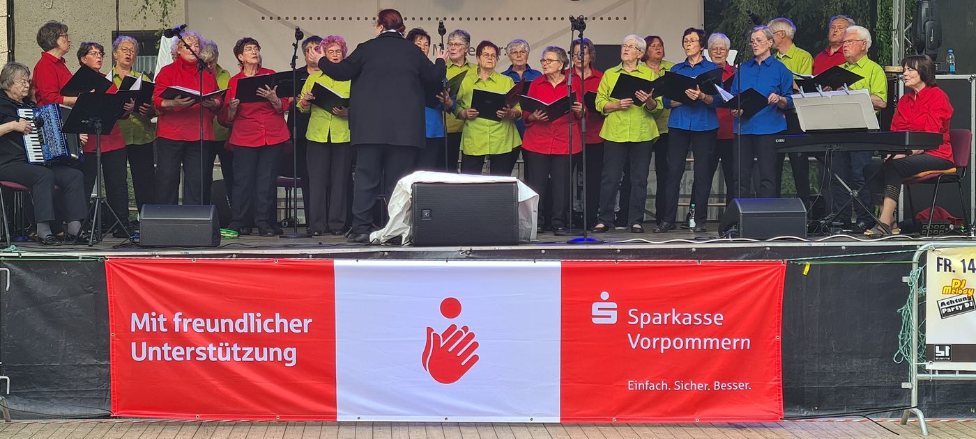 Ein Chor älterer Erwachsener singt unter der Leitung eines Dirigenten auf einer Freilichtbühne. Die Mitglieder tragen rote, lindgrüne und blaue Hemden. An beiden Enden sitzen ein Pianist und ein Akkordeonspieler. Vorne wirbt ein rot-weißes Banner für die Sparkasse Vorpommern.