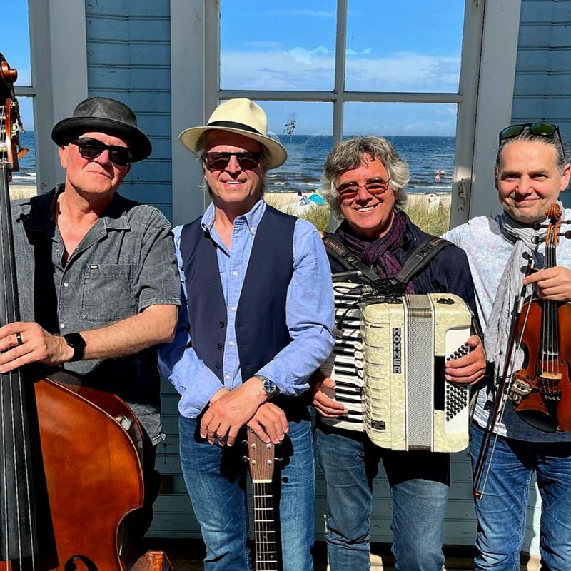 Vier Männer stehen zusammen, jeder hält ein Musikinstrument (Kontrabass, Gitarre, Akkordeon und Geige) und lächelt. Sie stehen vor einem hellblauen Holzgebäude mit großen Fenstern, die an einem sonnigen Tag den Blick auf das Meer freigeben.