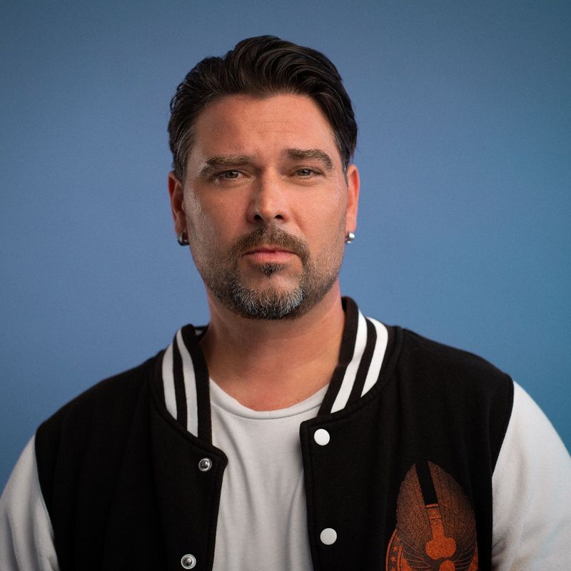 Ein Mann mit kurzen dunklen Haaren und gestutztem Bart, der eine schwarz-weiße Sportjacke über einem weißen T-Shirt trägt, steht vor einem einfarbig blauen Hintergrund und schaut mit neutralem Blick direkt in die Kamera.
