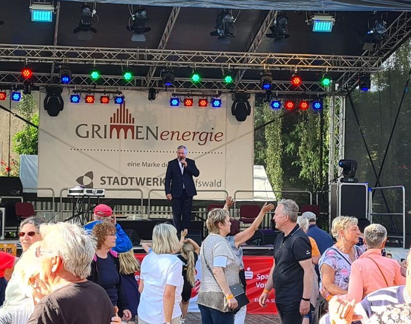 Eine Menschenmenge beobachtet einen Mann im Anzug, der auf einer Freilichtbühne mit bunten Lichtern auftritt. Im Hintergrund ist das GRIMMEnergie-Logo zu sehen. Vor der Bühne stehen Menschen und unterhalten sich im Sonnenlicht.