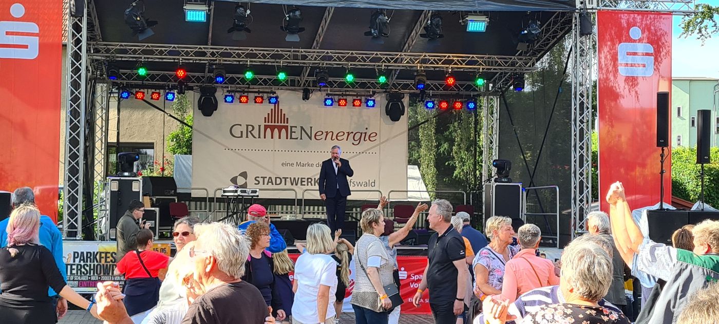 Eine Menschenmenge beobachtet einen Mann im Anzug, der auf einer Freilichtbühne mit bunten Lichtern auftritt. Im Hintergrund ist das GRIMMEnergie-Logo zu sehen. Vor der Bühne stehen Menschen und unterhalten sich im Sonnenlicht.