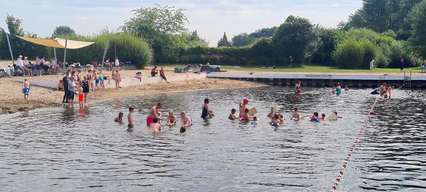 Im Schwimmbad tummeln sich Kinder und Jugendliche im Wasser. Einige lernen unter Anleitung von Trainern das Schwimmen. Im Hintergrund sind Liegewiesen und Sonnenterrassen zu sehen.